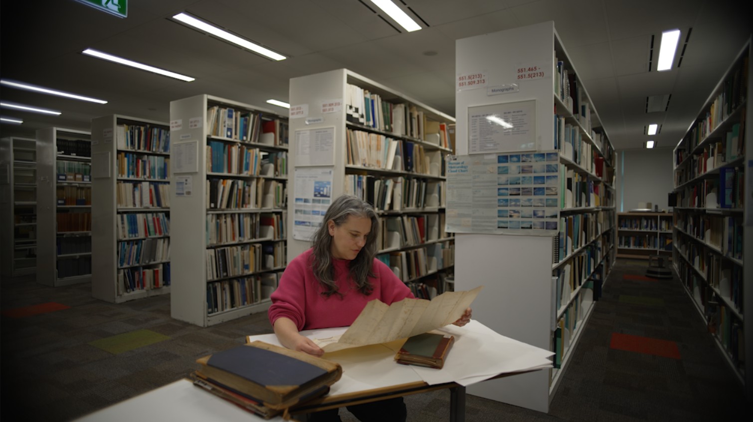 A woman wearing a pink jumper sits at a table looking at a document there are rows of books behind her.