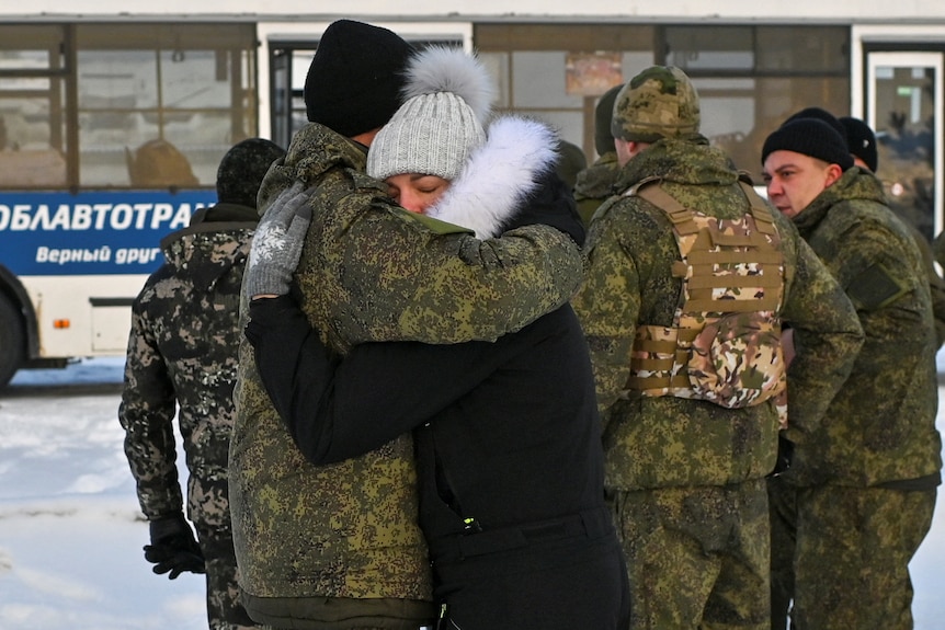 A Russian reservist in military uniform hugs a woman in a black coat, in front of other reservists and a bus.