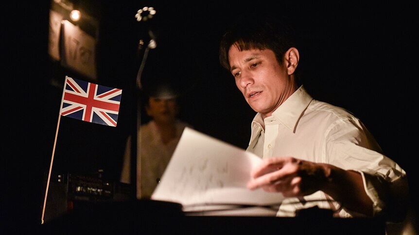 A man in white button up business shirt looks through papers on a desk with a small Union Jack flag in dimly lit room.