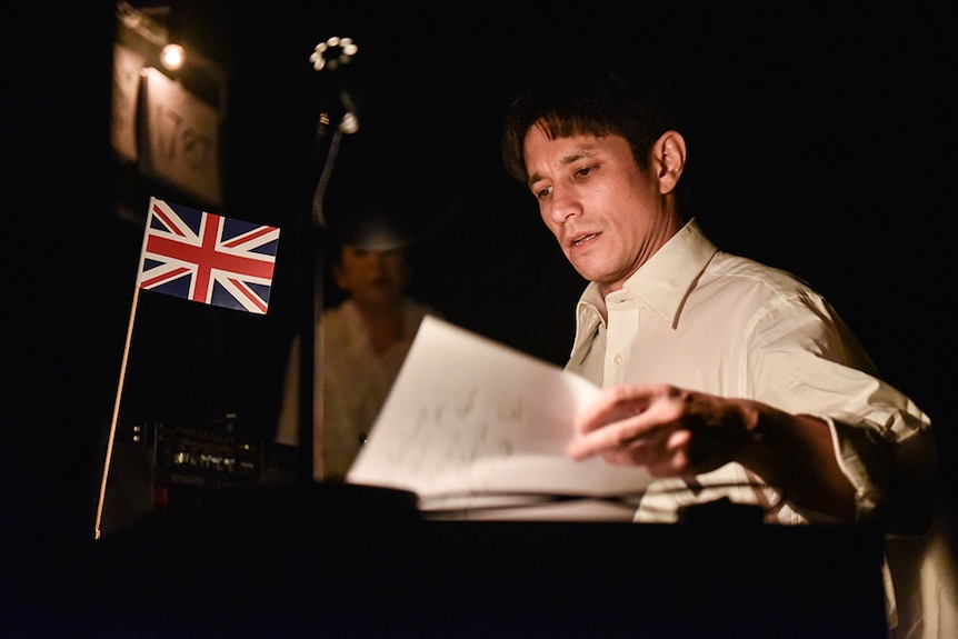 A man in white button up business shirt looks through papers on a desk with a small Union Jack flag in dimly lit room.