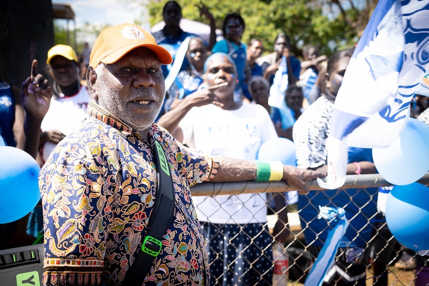 An Indigenous elder looks at the camera, leaning against a fence, with football supporters behind him