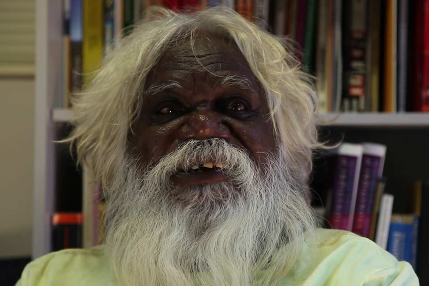 A close-up of an Aboriginal man with white hair and a beard, sitting in front of a bookshelf.