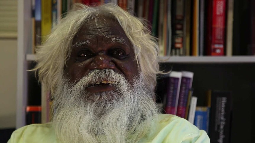 A close-up of an Aboriginal man with white hair and a beard, sitting in front of a bookshelf.