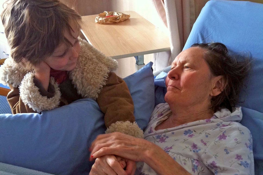 A woman lies in a nursing care bed while holding the hand of a young boy who has his hand on his face