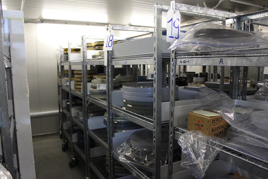 A white-walled room filled with metal racks stacked with film reels inside plastic and metal tins.