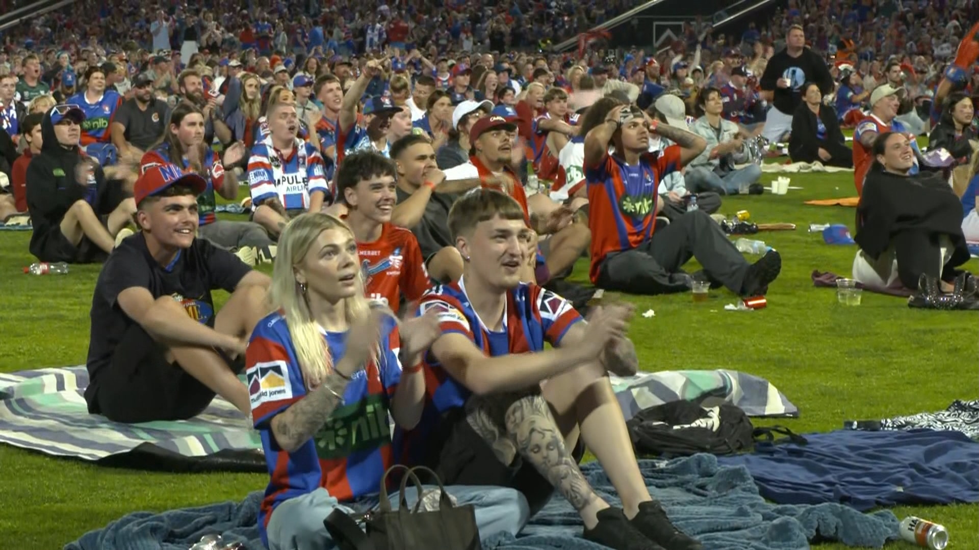 Scores of fans in red and blue jerseys sitting on grass in a stadium.