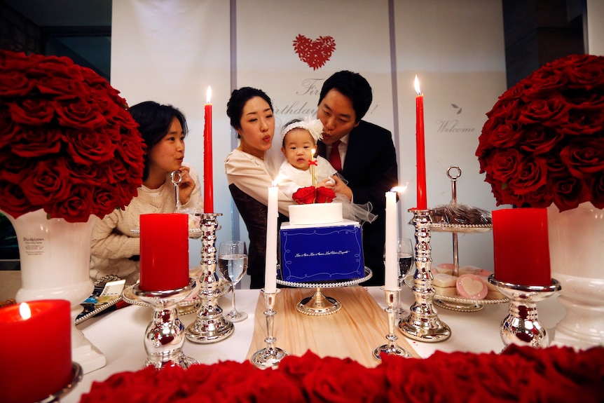 a man and woman hold an infant up to a birthday cake as they go to blow out the candles on a birthday cake on a decorated table