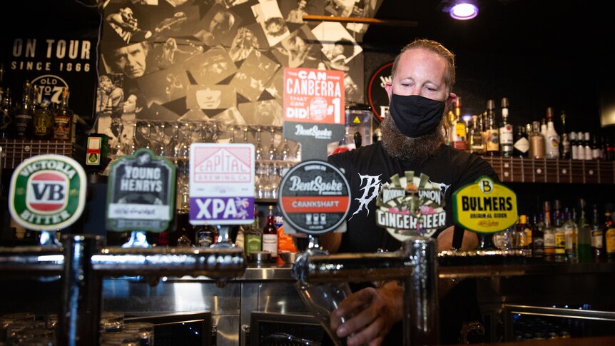 A man in a black face mask pours beer from a bar tap.