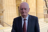 A bald man wearing a suit and tie outside a building with large brown columns