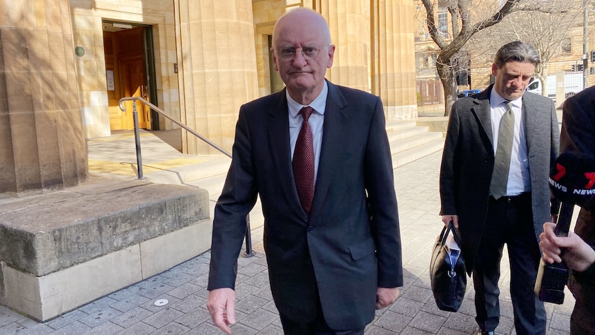 A bald man wearing a suit and tie outside a building with large brown columns