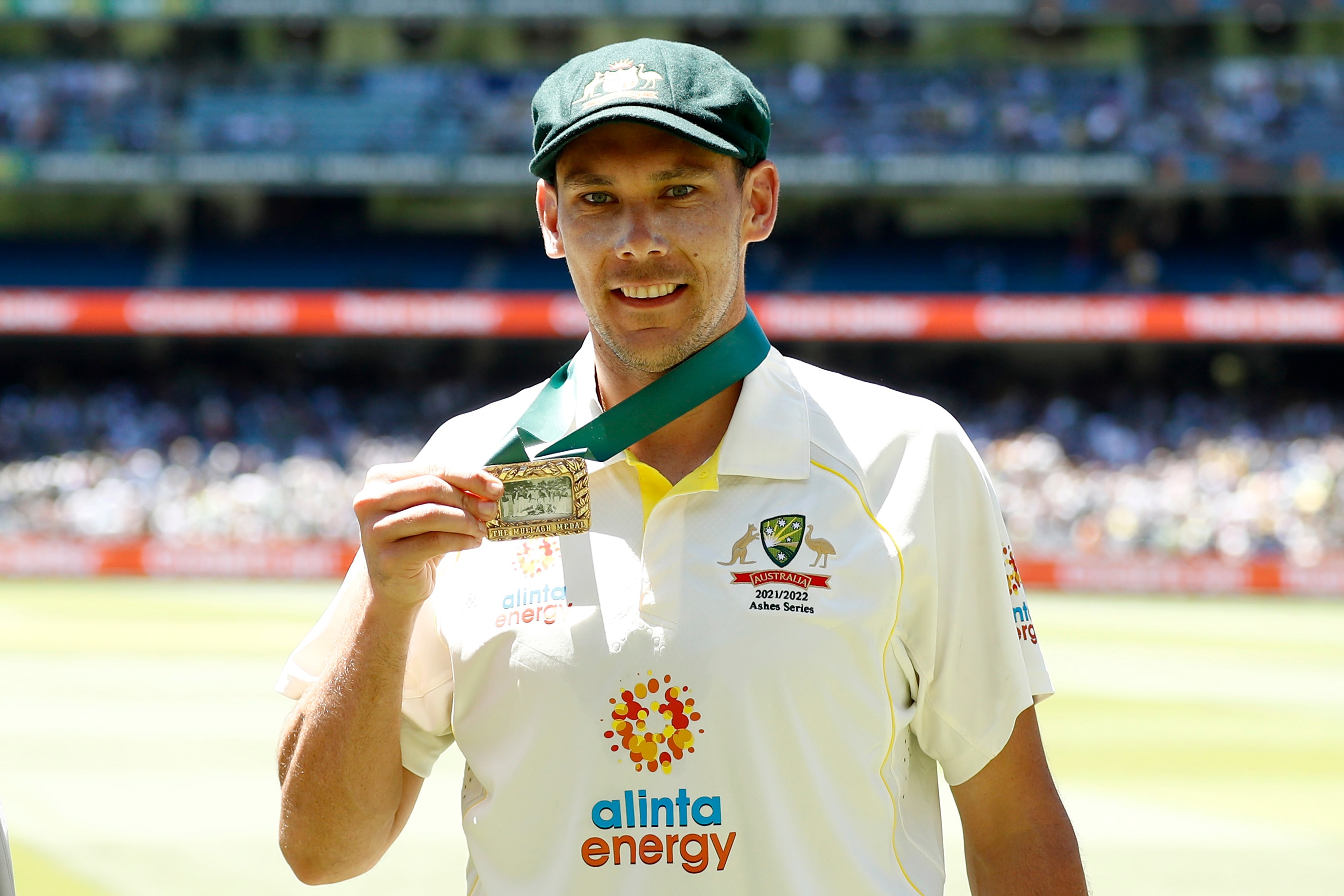 O jogador de críquete Scott Boland segura uma medalha no pescoço, sorrindo em frente às arquibancadas do Melbourne Cricket Ground.
