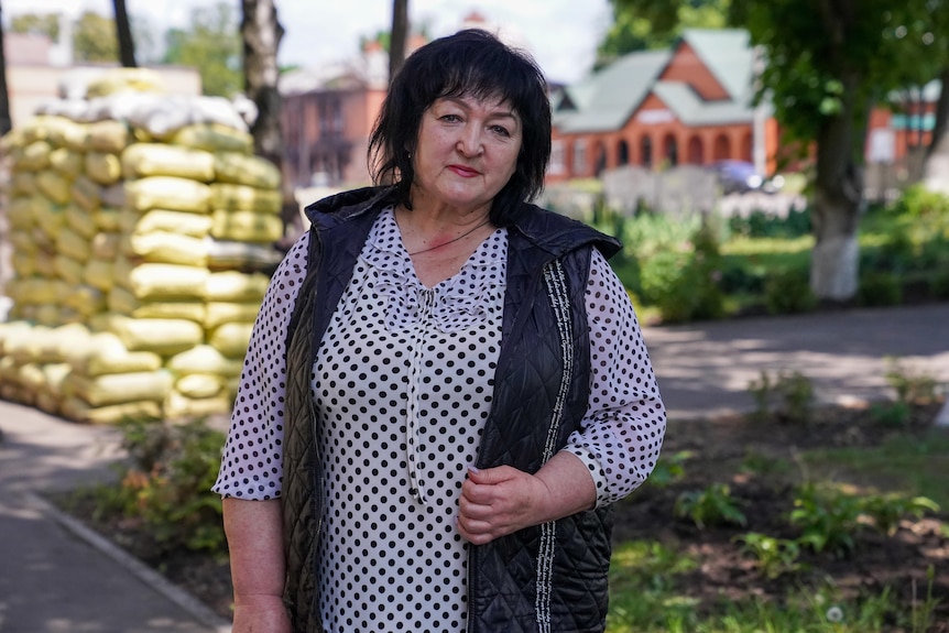 A woman with cropped black hair looks at the camera. Behind her sits an enormous pile of sandbags.