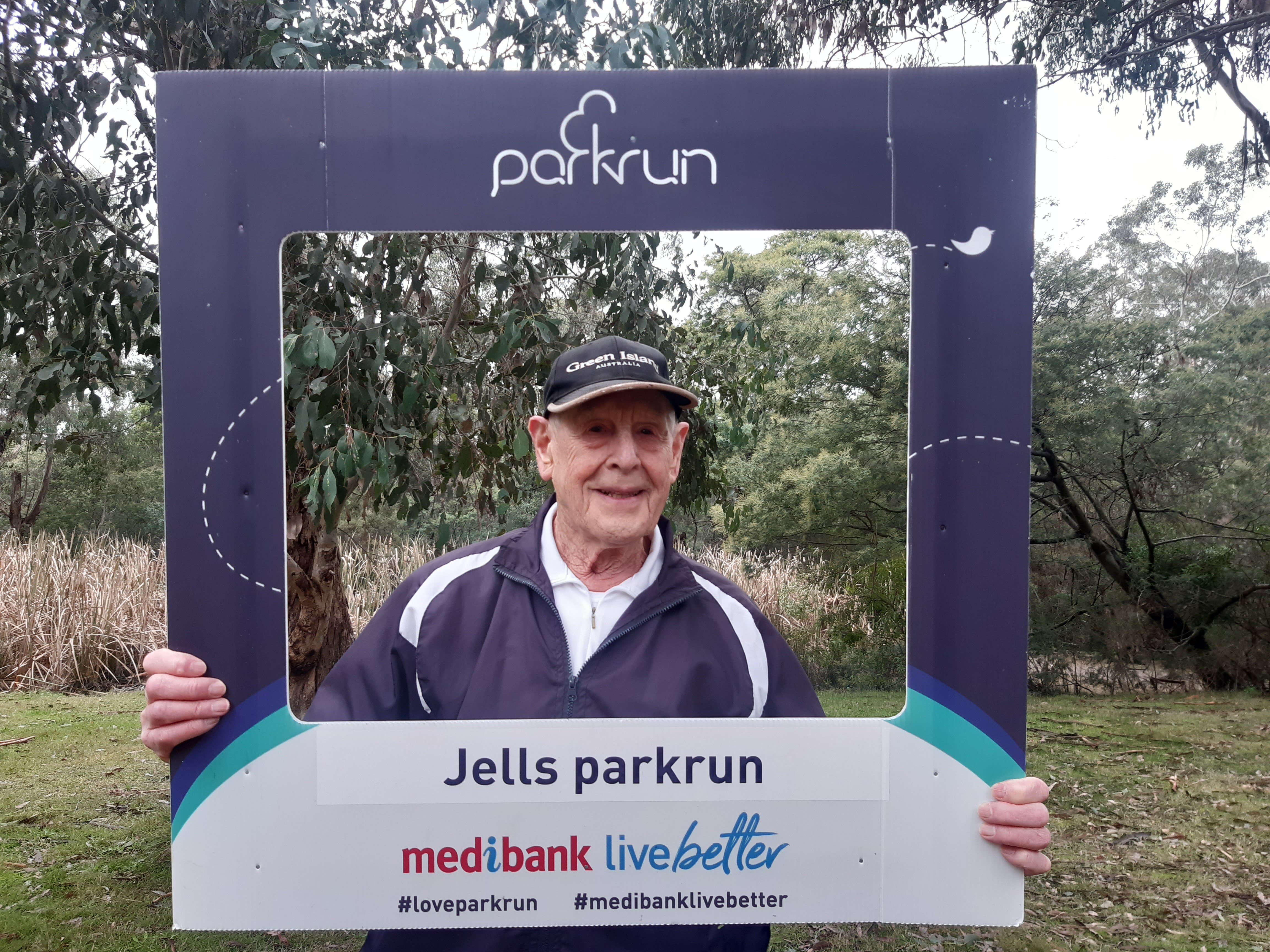 93-year-old Bill Lamont holds a parkrun cut-out frame and smiles.
