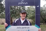 93-year-old Bill Lamont holds a parkrun cut-out frame and smiles.