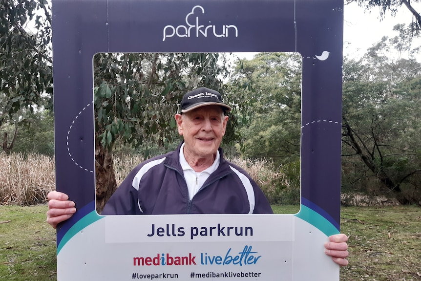 93-year-old Bill Lamont holds a parkrun cut-out frame and smiles.