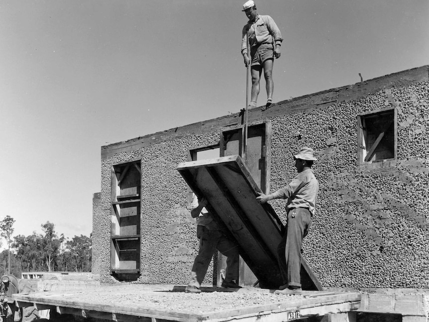 Three people lift a wooden frame from a truck. One is standing on top of a semi-built wall of a house.