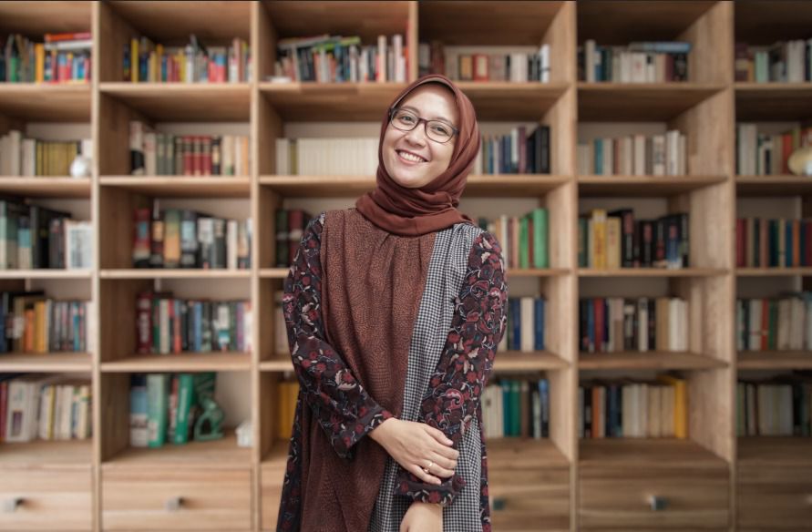 Una mujer con gafas y hijab cruza la mano y sonríe a la cámara.