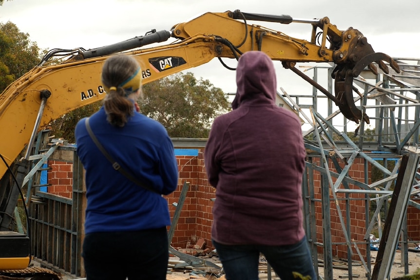 Two women with their backs turned with a digger demolishing a home in the background