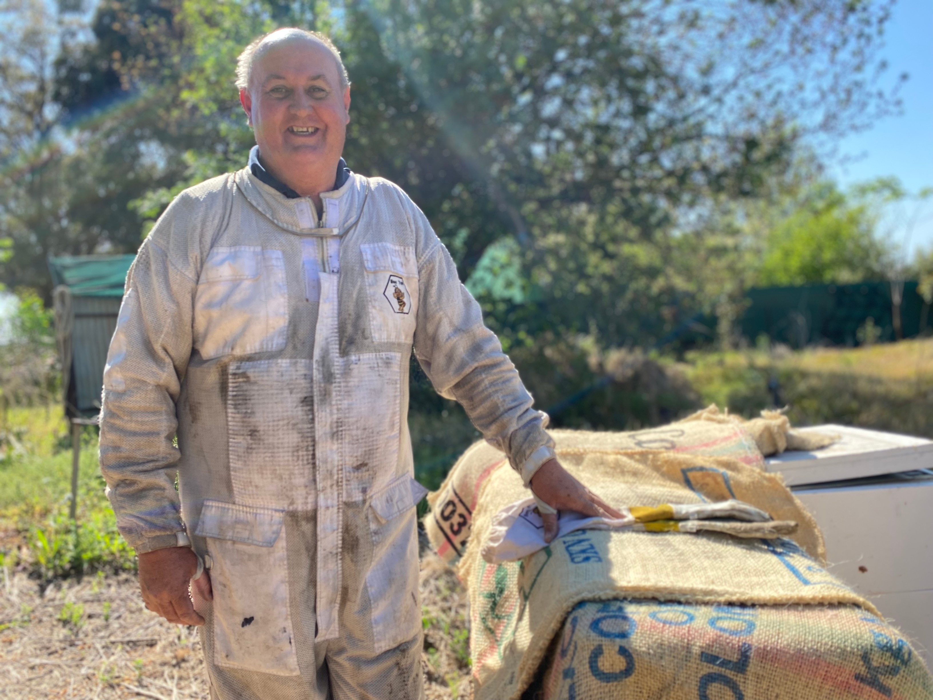 A tall man in a white beekeeping suit zipped up to his neck smiling and leaning on a box.