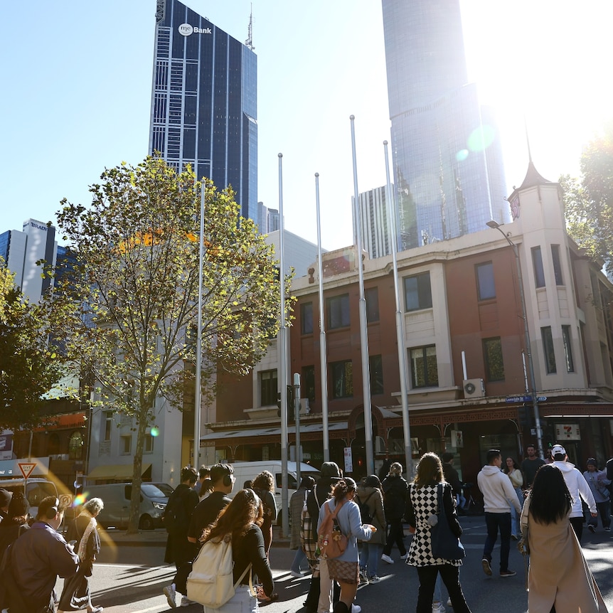 A crowd of people walking in a city with high rise buildings in the background on a sunny day.