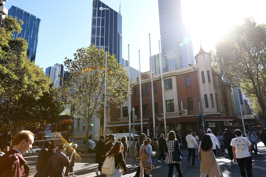 A crowd of people walking in a city with high rise buildings in the background on a sunny day.