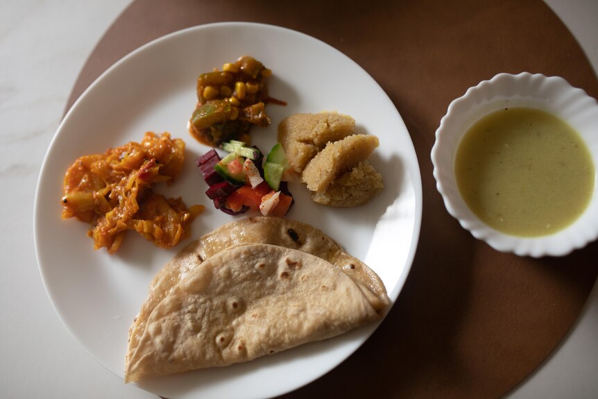 A white plate with Indian food, three vegetables, one dessert, salad, Indian bread and a bowl of yellow sauce.