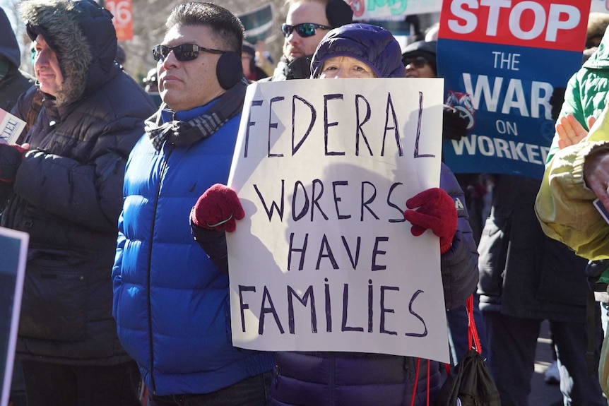 Workers stand in the street protesting with various placards. One woman holds a sign reading: Federal workers have families.