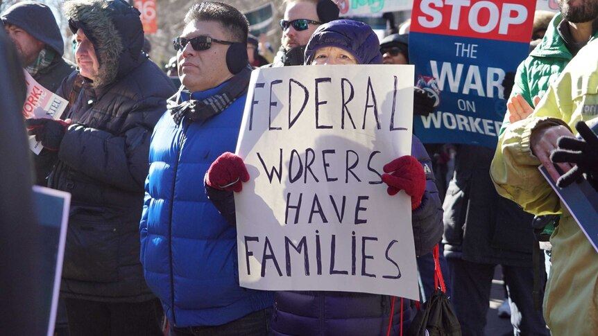 Workers stand in the street protesting with various placards. One woman holds a sign reading: Federal workers have families.