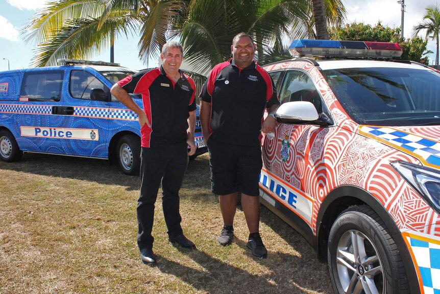 Two men stand beside two police cars decorated with Aboriginal art.