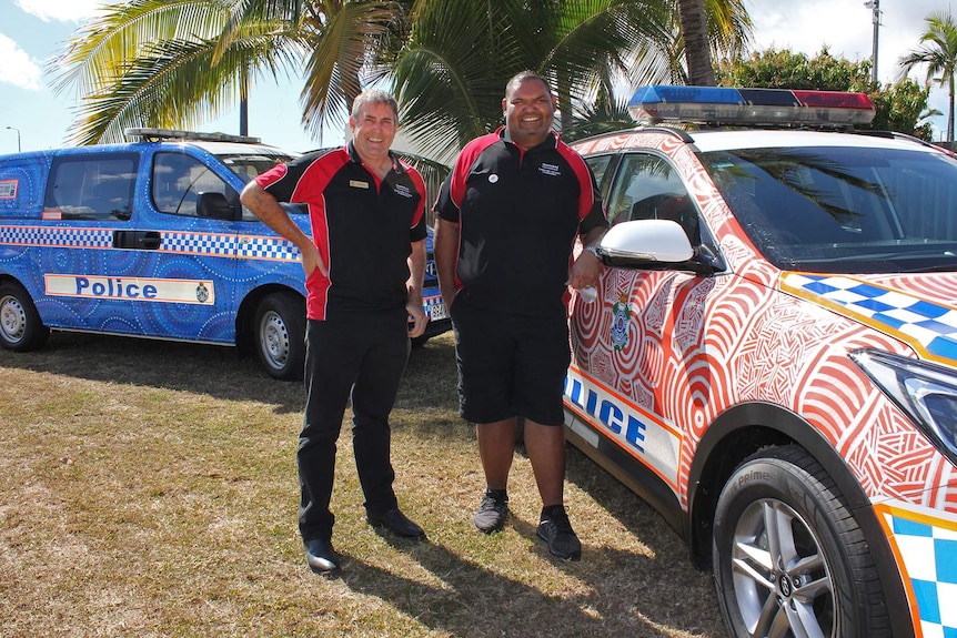 Two men stand beside two police cars decorated with Aboriginal art.