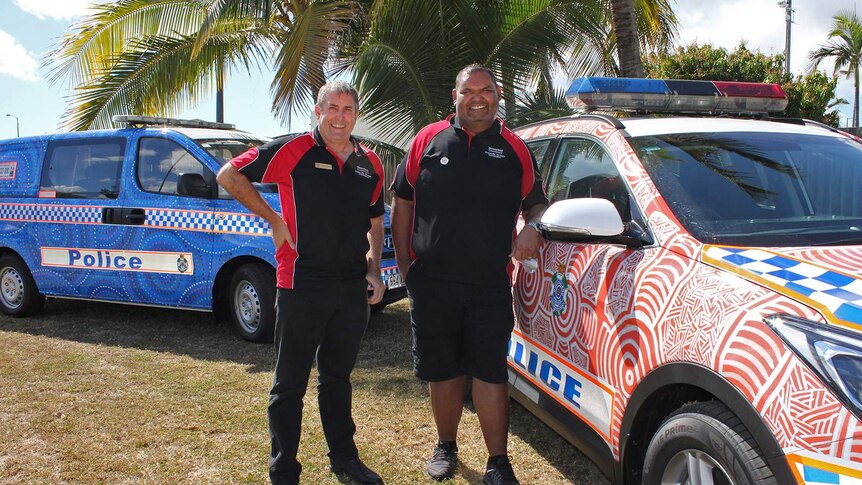 Two men stand beside two police cars decorated with Aboriginal art.