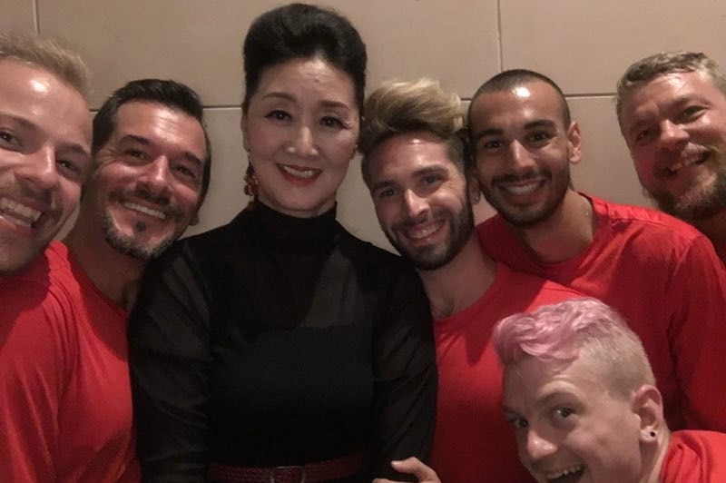 A woman wearing a black turtle neck smiles while surrounded by six men in red T-shirts.