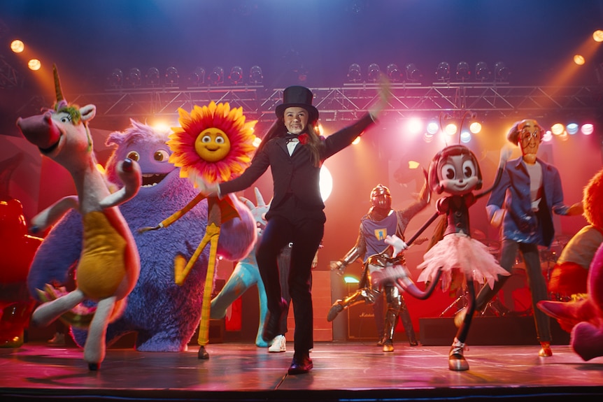 A young girl in a top hat dances on stage alongside a lot of animated creatures and monsters