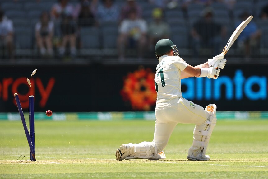 Bails come off stumps as a cricket ball hits them behind Australia batter David Warner, who is completing a stroke.