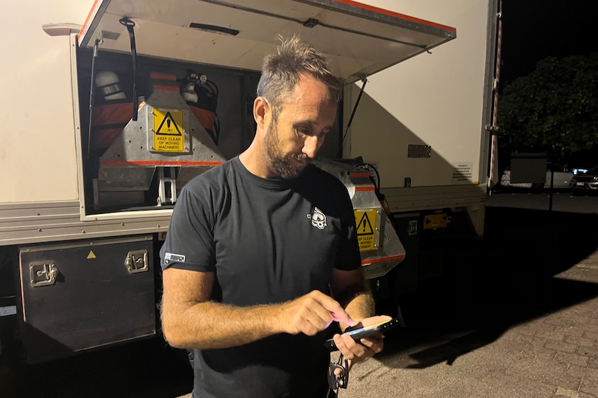 A middle-aged man in black T-shirt looks down at his phone next to back of trucks in evening.