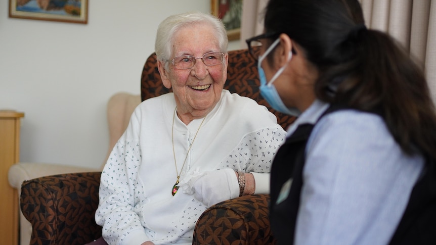 A woman with white hair and a white jumper sits on a couch smiling, facing a woman wearing a mask who has her back to the camera