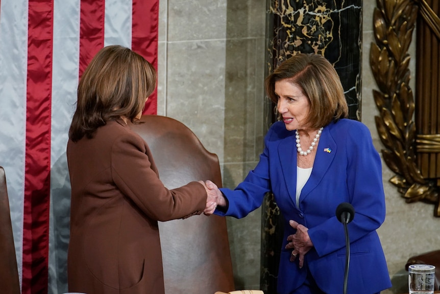 Nancy Pelosi in a blue suits and pearls shakes hands with Kamala Harris in a brown suit