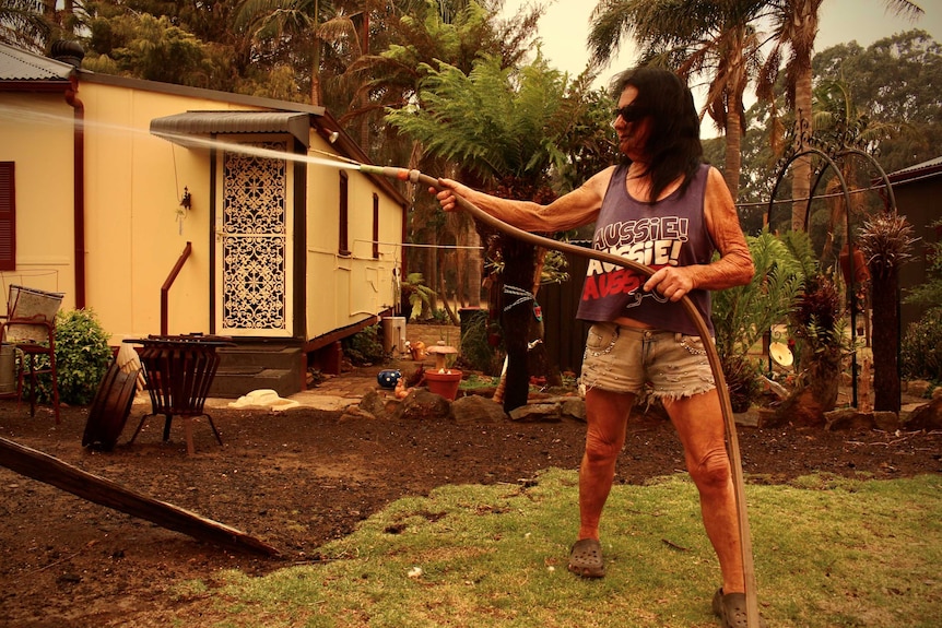 A woman wearing shorts and a singlet top points a hose as she stands in front of a house.