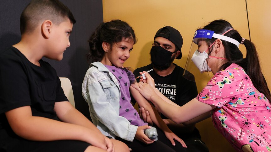 A boy and a man looks on as a young woman in pink shirt, face shield, face mask injects a girl.
