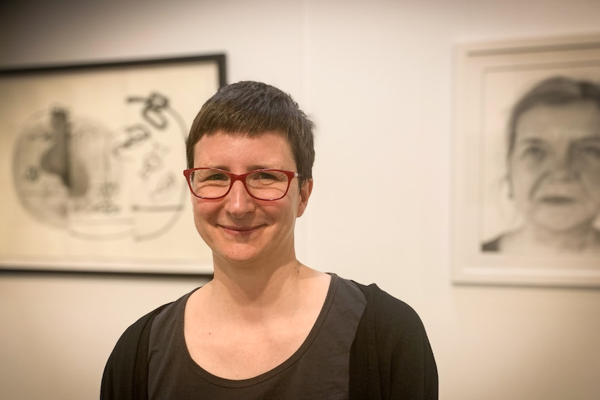 Photo of a woman with short hair and glasses in front of two portraits