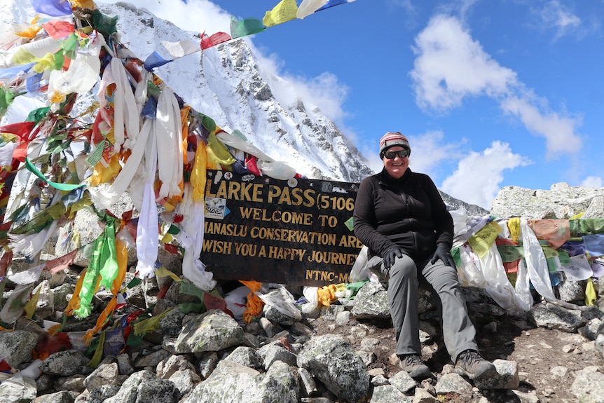 A woman sits on stones in front of a sign marking a mountain trail. 