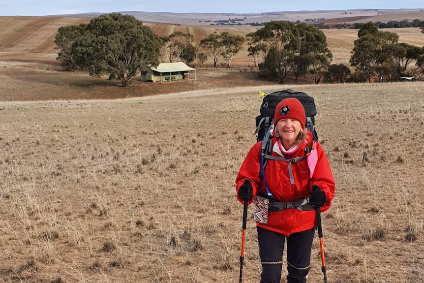 A woman in hiking gear holding walking poles stands on a grassy hill with a small hut in the background