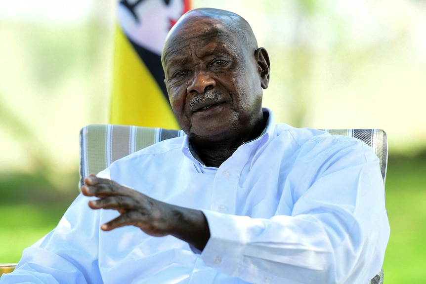 An older Ugandan man in a light blue business shirt gestures as he speaks in front of a blurred background of green plants.