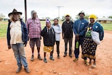 Seven Martu people stand outside with houses in the background at the Parnngurr Indigenous community.