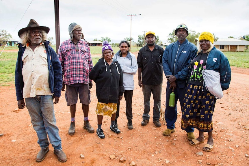 Seven Martu people stand outside with houses in the background at the Parnngurr Indigenous community.