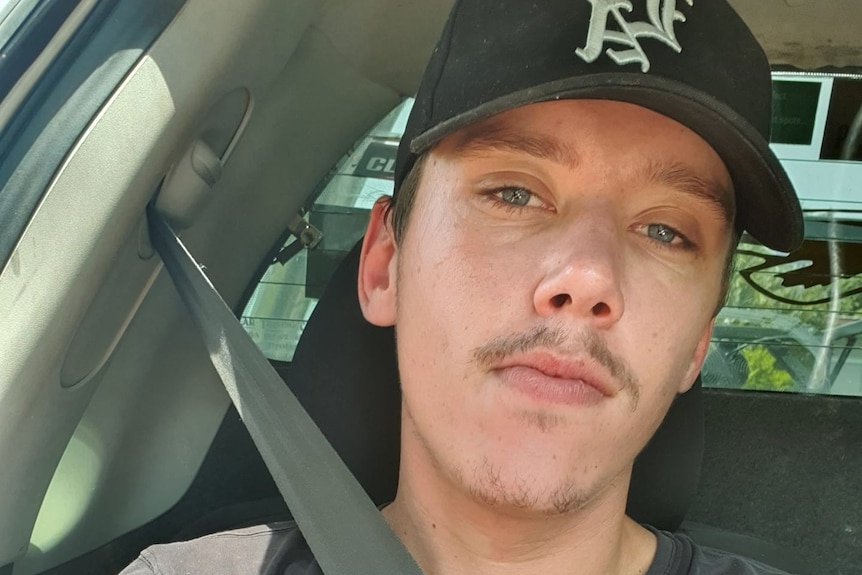 A young man with a moustache wearing a hat while sitting in the back seat of a car.