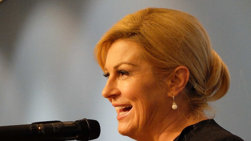 A side profile of a woman with blonde, pinned back hair smiling as she speaks into a microphone, with a soft grey background.