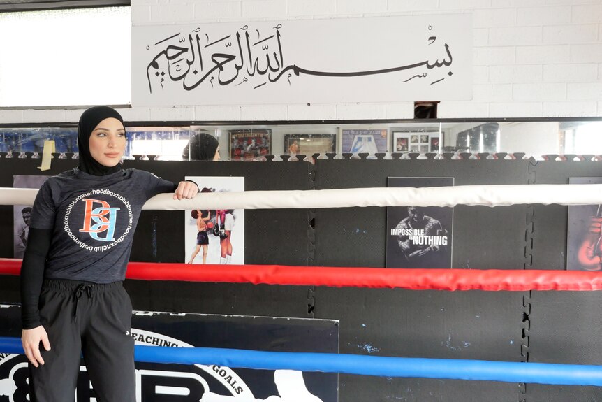Una boxeadora que lleva un hiyab se apoya contra las cuerdas, se muestra un extracto del Corán sobre ella.