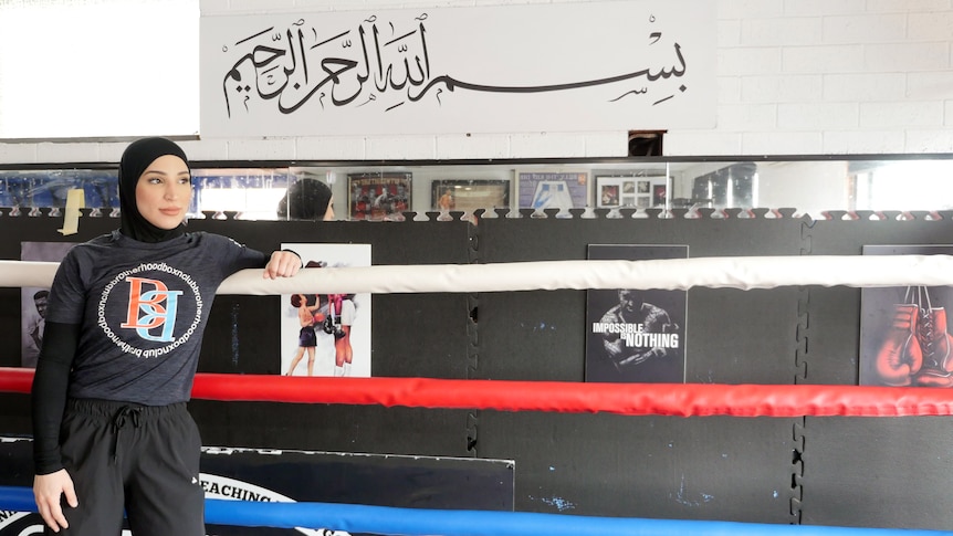 A female boxer wearing a headscarf leans against the ropes, an excerpt from the Quran is displayed above her.