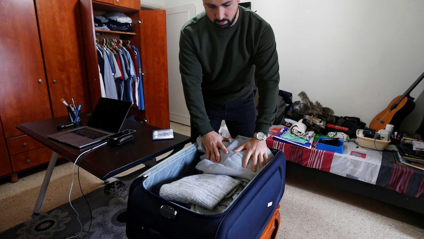 A man in a dark jumper packs a navy blue suitcase full of clothes